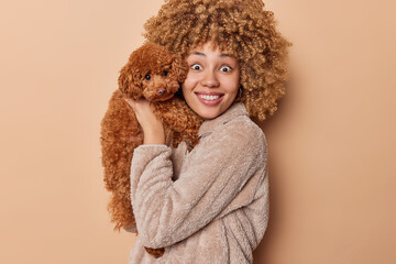 Portrait of pretty curly haired young woman with poodle puppy near face hugs new lovely member of family looks with eyes full of interest dressed in winter coat isolated over brown background.