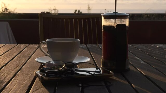 Caf&eacute; reci&eacute;n hecho en cafetera de &eacute;mbolo con bonita luz de amanecer y m&oacute;vil y gafas de lectura. Entra en el plano una taza de caf&eacute;.