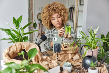 Professional curly female gardener records gardening videos teaches how to grow plants holds bulb plant with roots uses modern devices takes care of potted flowers poses indoor. Broadcasting