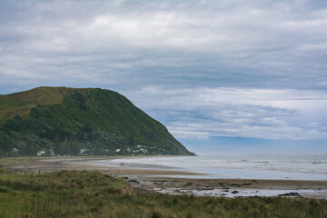 Distand couple walking along vast ocean beach surrounded by rugged mountains. Heavy clouds and thick mist over riptide. Makorori beach, Gisborne, North Island, New Zealand