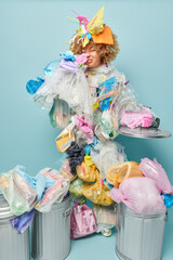 Displeased curly haired woman covers nose smells unpleasant odor from rubbish collects litter surrounded by different bins coveres with plastic wastes isolated over blue wall. Environmental pollution