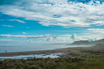 Lone surfer walking with a white board along water edge at Makorori Beach, mist rising over water obscuring distant mountains. Gisborne, New Zealand