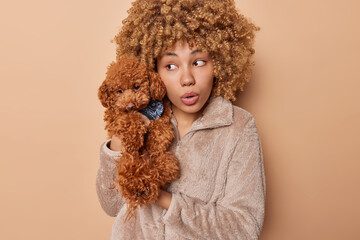 Portrait of surprised woman looks with great wonder aside holds pedigree puppy small poodle dog going to have walk in park dressed in coat isolated over brown background. Stunned female pet owner