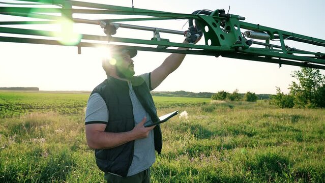 Male farmer engineer stand next to tractor and uses mobile application on digital tablet PC. Mechanic online optimizes work of small businesses in production of products and repairs machinery.