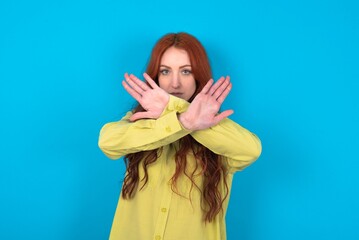 young woman wearing green shirt over blue background has rejection expression crossing arms and...