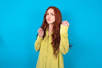 young woman wearing green shirt over blue background Ready to fight with fist defense gesture, angry and upset face, afraid of problem.