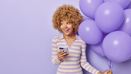 Happy birthday girl dressed in casual striped jumper uses mobile phone receives sms of congratulation holds bunch of inflated balloons focused aside isolated over purple background copy space for text