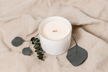 Candle in glass jar with wooden wick on beige linen cloth with eucalyptus leaves. Aesthetic home decoration, bohemian style