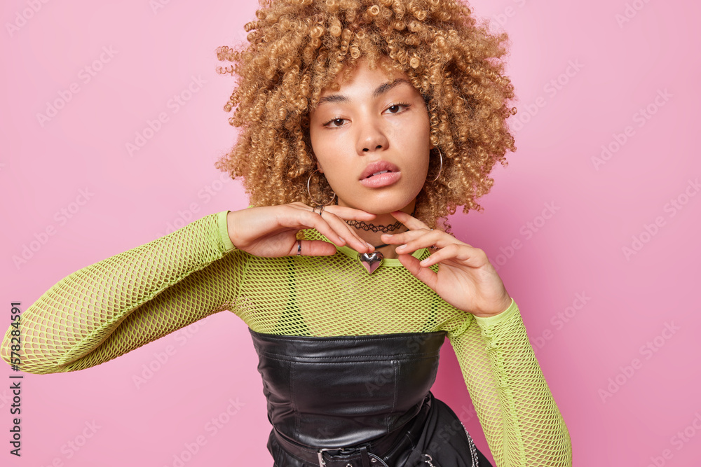 Wall mural Portrait of serious curly haired woman touches jawline gently focused directly at camera dressed in fashionable clothes has healthy skin calm expression isolated over pink studio background.