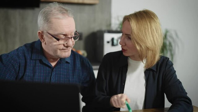 An elderly retired man and his adult daughter sit at home and work with paper bills or tax forms. A woman helps her father work with a laptop.