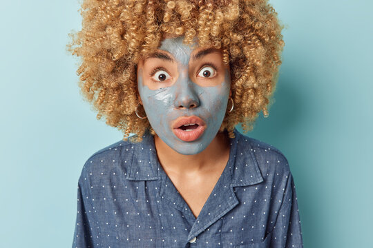 Portrait of scared curly haired woman applies nourishing clay mask to reduce fine lines stares impressed with widely opened eyes and mouth dressed in nightwear isolated over blue background.