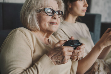Cute emotional senior woman and daughter playing videogame holding gamepad controllers sitting on...