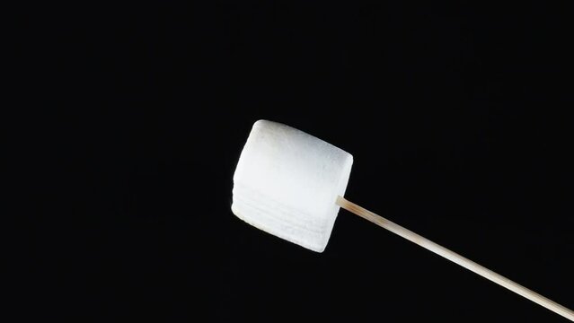 Marshmallow rotates on a stick and browns to a caramel crust, Close-up, isolated object on a black background.