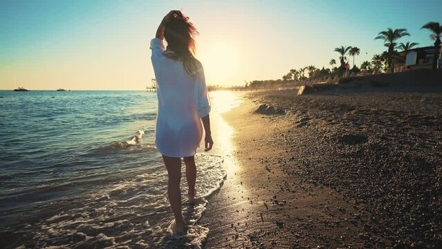 Happy woman silhouette walks beach blue water of ocean. Young woman walking on beach under sunset light. Summer tropical mood. Summer holidays, lifestyle, vacation at sea.