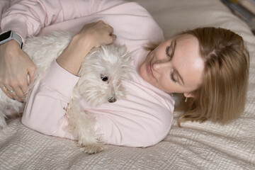 Woman hugging little white lap dog while laying on bed together
