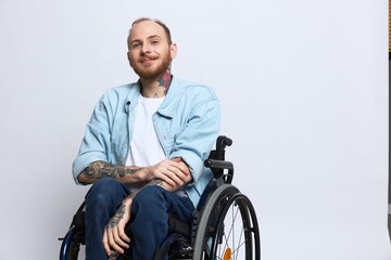 A man in a wheelchair smile, copy space, with tattoos on his arms sits on a gray studio background,...