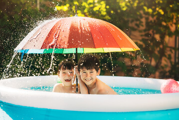 Two little children have fun in the inflatable pool at home outside. Kids hide from the falling splash of water under a colorful umbrella. Water entertainment outdoors.