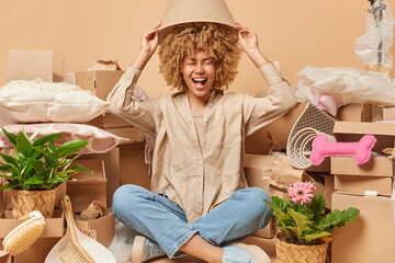 Cheerful young female renter sits crossed legs covers head with lampshade going to move and unpack carton boxes wears shirt jeans and sportshoes. First dwelling relocation and mortgage concept