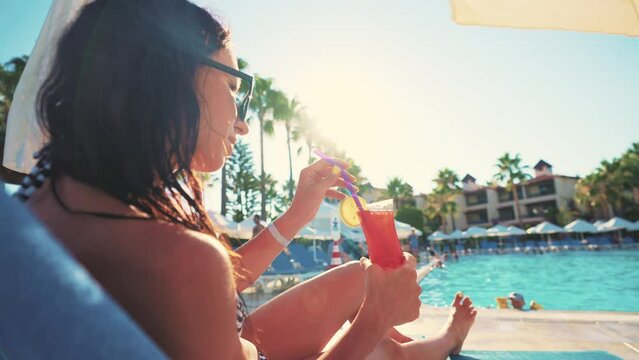 Fresh cocktails in hands. Young woman have fun in swimming pool at daytime. Cooling cocktail with ice from table and drinks it through straw. Resort and relaxing on territory spa hotel. Summer.
