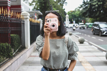 Attractive young girl in casual style taking picture by pink instax camera at the road side