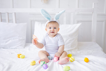 cute baby boy with bunny ears and colorful eggs on a white bed at home playing or eating eggs, little blonde baby, happy easter concept