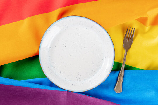Gay dinner party invitation. A place at a restaurant table with a plate and a fork, overhead flat lay shot on the LGBT rainbow flag texture