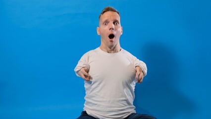 a young man with disabilities on a blue background is afraid and shocked by surprise and astonished...