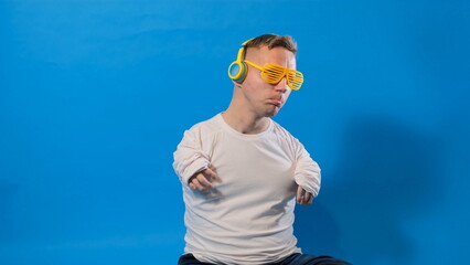 a man with disabilities in stylish glasses and yellow headphones listens to music and dances on a blue background in the studio.