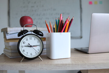 Concept to teacher's day. Pens, apple, pencils, books, alarm clock and laptop on the table, against the background of whiteboard. Back to school, copy space.