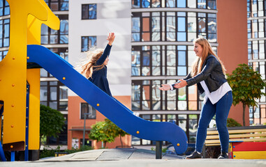 Cheerful girl raising hands up while getting off slide, mom waiting below at modern courtyard of...