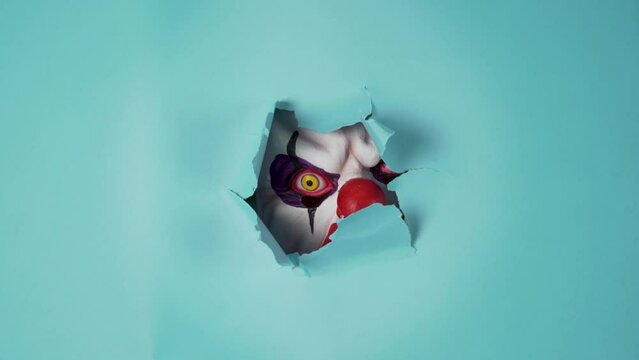 creepy clown stock photo is sure to give you goosebumps, featuring a haunting blue background and a sinister figure peering through a paper hole. Perfect for Halloween-themed promotions or horror