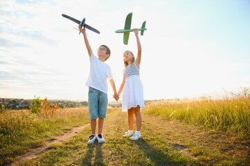 Obraz premium Running boy and girl holding two green and blue airplanes toy in the field during summer sunny day
