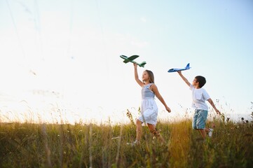 Running boy and girl holding two green and blue airplanes toy in the field during summer sunny day