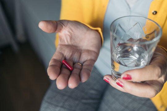 Cropped image of an unrecognizable older woman in yellow sweater holding a glass of water in one hand and a red pill in another hand to take her medication sitting on her bed.