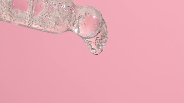 Drops of a liquid transparent product for care and hygiene are dripping from a glass pipette on a pink background. Dropper with serum, gel with bubbles macro.