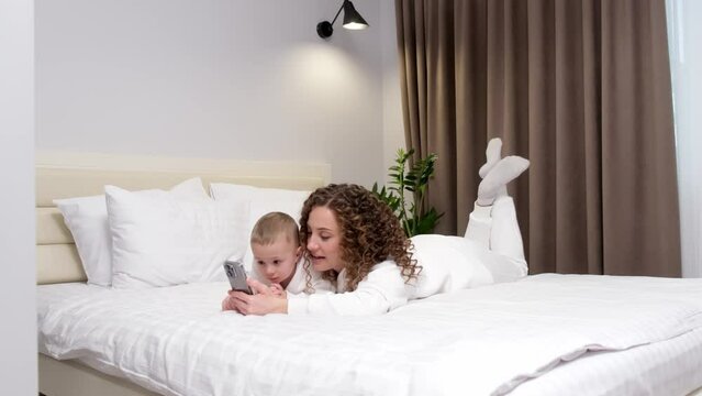 Mom and little 2-year-old son are lying on the bed, looking at a mobile phone, laughing, watching cartoons, making video calls, having fun on the Internet. Two generation family use new app, modern te