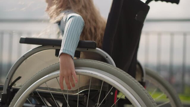Hands of little girl with loose curly hair spinning large wheels of medical vehicle. Child with disability learns to move in wheelchair on street closeup