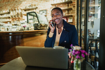 Portrait of a smiling young woman using laptop and talking on mobile phone in coffeeshop