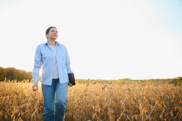 Naklejka na ściany i meble Young farmer woman standing in soy field in summer time, holding tablet and monitoring crop growth