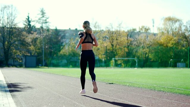 Rear view. An adult sports female runs on the track of the urban city stadium. A beautiful fit woman in a tracksuit makes her daily jog on the street. A healthy young athlete is busy with training