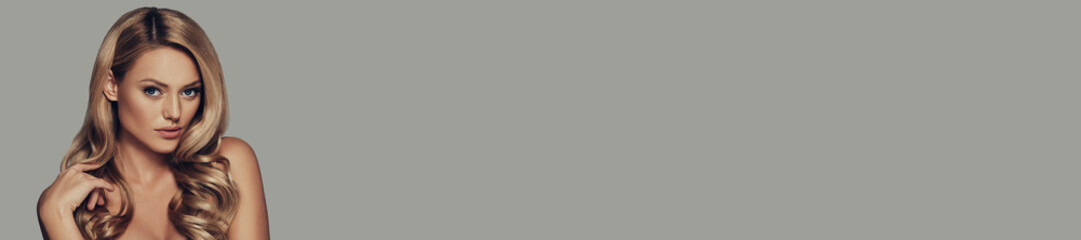 Fototapeta premium Gorgeous young woman with long and waving blonde hair looking at camera against grey background