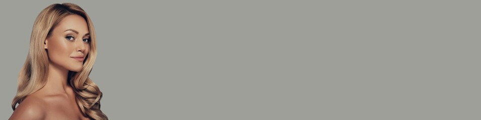 Fototapeta premium Attractive young woman with long and waving blonde hair looking at camera against grey background
