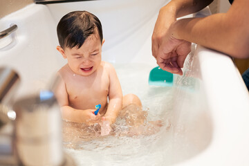 A multiethnic Asian male infant crying in the bathtub while his father is giving him a bath with warm or lukewarm water. The baby is feeling unwell and holding a toothbrush on his hand.