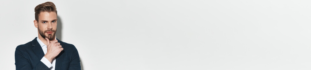 Handsome young man in suit holding hand on chin while standing against grey background