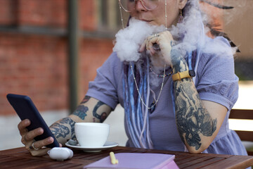 Close-up of young woman with e-cigarette sitting by table in outdoor cafe, blowing steam out of her...