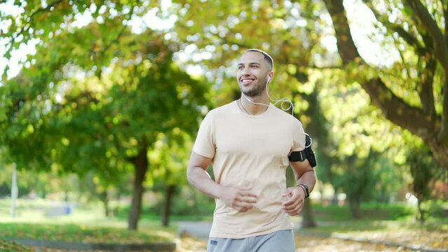 The young runner of mixed race with headphones and a smartphone jogging in an urban city park. A beautiful man in a tracksuit likes to run outdoors under his favorite music. Нealthy lifestyle concept
