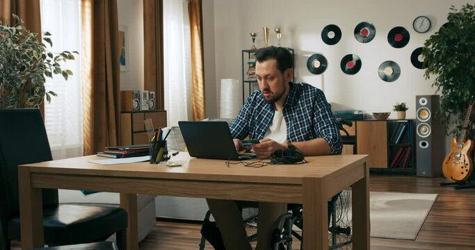 Person with disabilities beard man in wheelchair works from home online. Delighted happy man finishes work freelancer concept. Person closing laptop putting on table credit card easy payment.