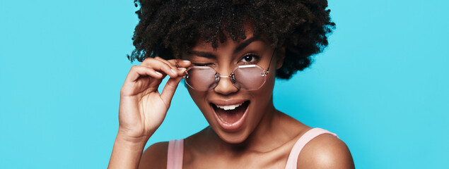 Attractive young African woman adjusting eyeglasses and winking against blue background