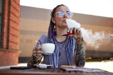 Young woman with tattoos on arms relaxing in outdoor cafe while having cup of coffee and blowing...
