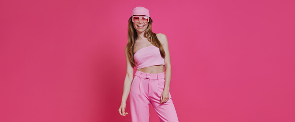 Beautiful young woman in trendy clothing standing against pink background